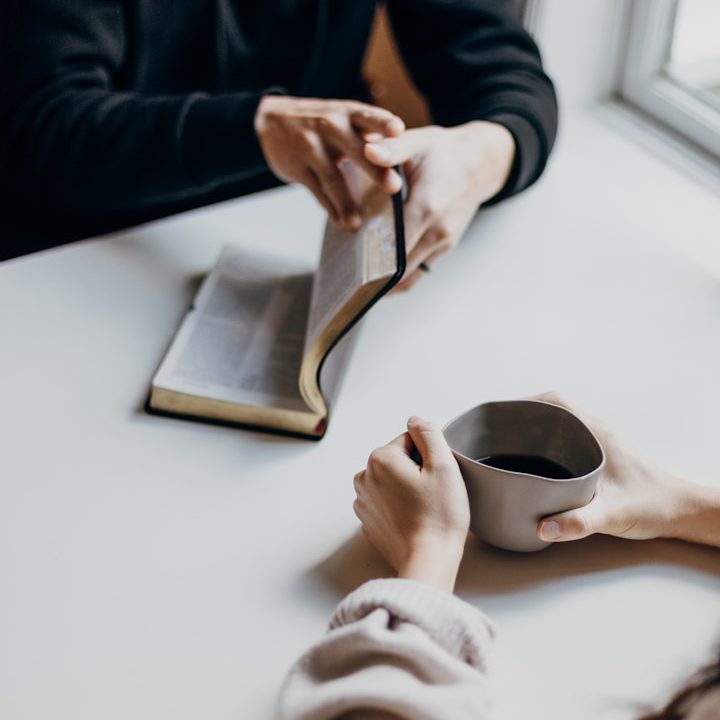 Zwei Personen sitzen sich an einem Tisch gegenüber, eine hält eine Tasse, die andere ein aufgeschlagenes Buch.