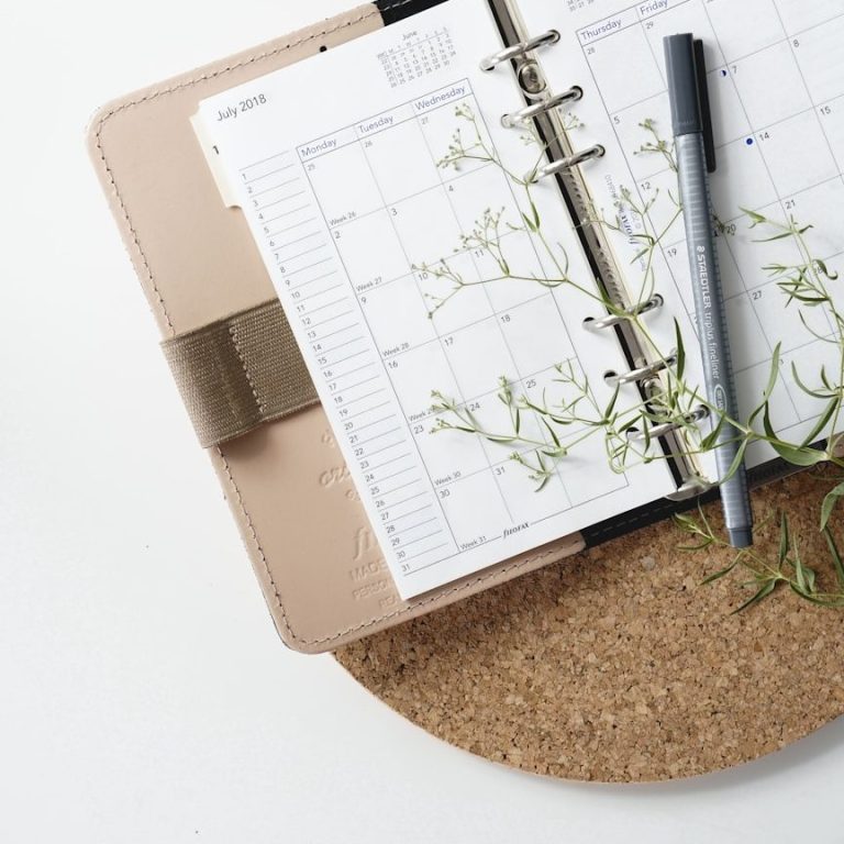 Ein offenes Notizbuch mit Kalender, einem Stift und edlen Pflanzenzweigen auf einem Untersetzer im Hochzeitslook.