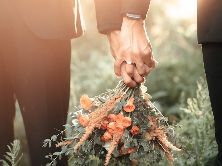Hände von zwei Personen bei ihrer freien Trauung, die einen Strauß mit orangefarbenen Blumen halten.